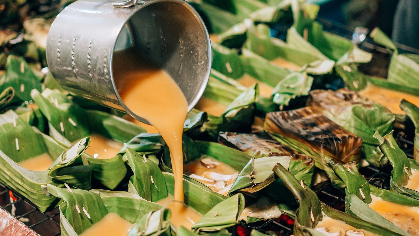 A yellow mixture poured from a metal jug into banana-leaf cups on a grill, at a street stall in Chiang Mai