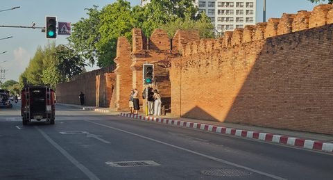 The inner face of the wall beside Tha Phae Gate, along Moon Muang Road, glowing in low afternoon sun, with a red songthaew and traffic lights at the crossing