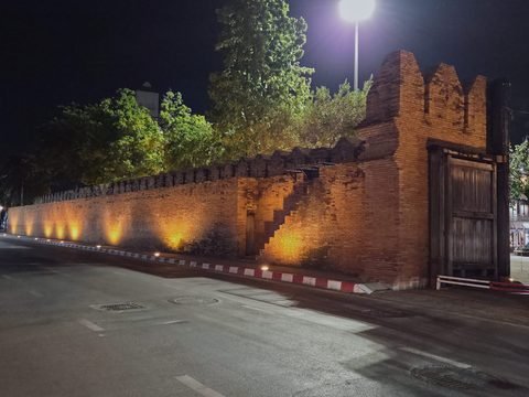The inner face of Tha Phae Gate at night from Moon Muang Road: floodlit brickwork stepped like a staircase climbs the wall beside a narrow doorway, with the back of the gate's wooden door on the right