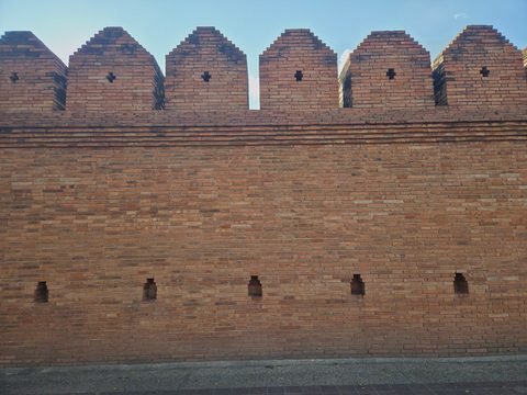 A straight-on view of the brick wall beside Tha Phae Gate: pointed merlons pierced with small cross-shaped loopholes along the top, a projecting band of brick below them, and a row of small holes near the base