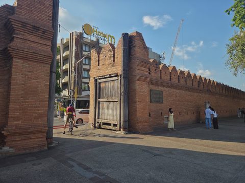The opening of Tha Phae Gate seen from the square: a cyclist rides through between the two brick towers, past the wooden door and its tall timber post, with a modern hotel rising behind the wall