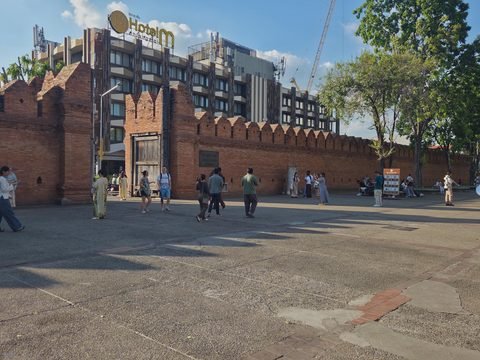 Tha Phae Gate and its square in the late afternoon: visitors walk across the paving in front of the crenellated wall, long tree shadows stretch towards the camera, and Hotel M stands behind the gate