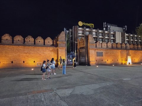 Tha Phae Gate at night from the square: the wall glows orange under lamps set in the paving, small groups of visitors take photos, and the hotel behind is lit against a black sky