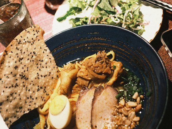 A bowl of khao soi with pork and its crisp fried noodles, a salad plate and drinks beside it