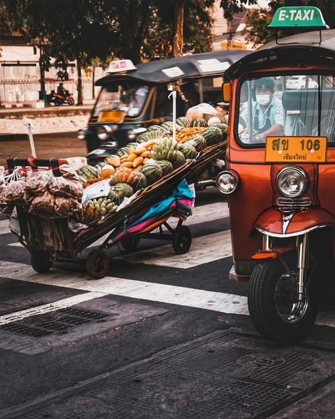 A handcart piled with watermelons, bananas and other fruit on a street crossing, beside two tuk-tuks in daylight