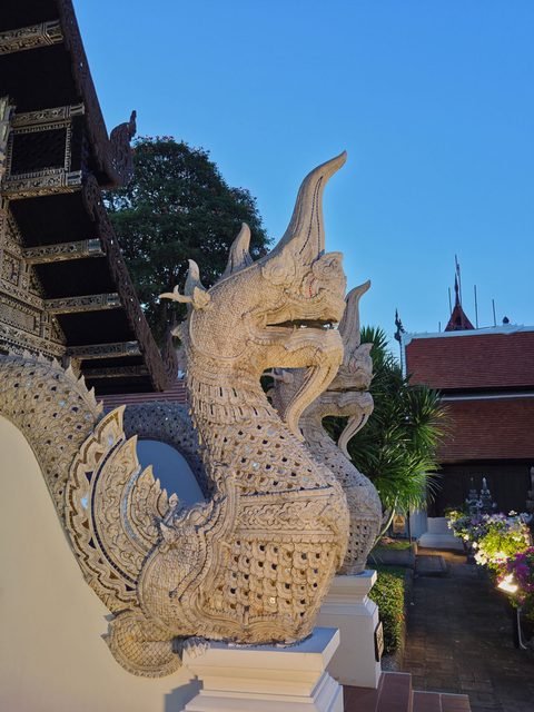 The stair of a carved teak hall at dusk: a white stucco naga rears up at the end of the balustrade, its crest curving back, with a makara at its foot