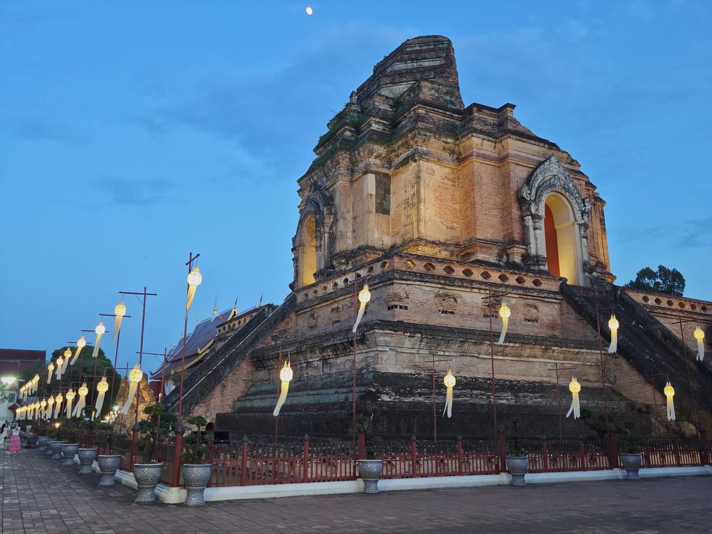 The great brick chedi of Wat Chedi Luang just after sunset, its broken top against a blue sky with the moon, white paper lanterns lit along the fence