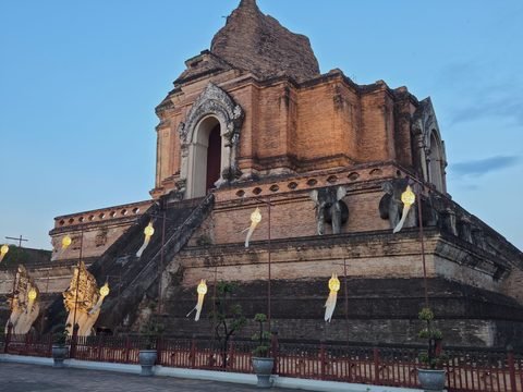 The chedi seen from a corner at dusk: two arched niches on the upper storey, elephants along the terrace, a steep stairway flanked by nagas, lanterns lit