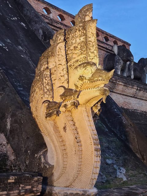 A floodlit stucco naga seen in profile, its main head with smaller heads below, two terrace elephants behind against the evening sky