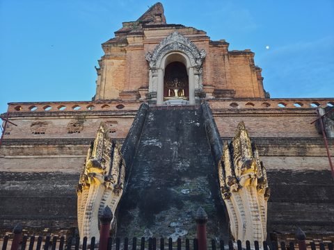 The chedi seen from the foot of a stairway at dusk: two floodlit nagas, the steep dark ramp, a lit niche with a seated gilded Buddha, and the moon in the sky