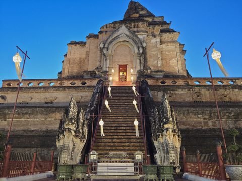 The east face of the chedi after sunset, seen head-on: a long stairway of real steps with a handrail between two stucco nagas, lanterns hanging over it, and at the top a small lit doorway in the niche
