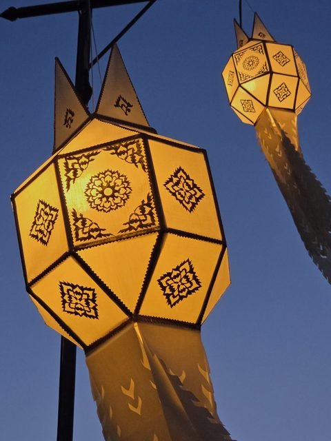 Two lit paper lanterns with cut-out patterns and long fringed tails, hanging from a pole against a deep blue sky