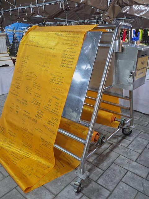 A long roll of yellow patterned cloth covered in handwritten names and wishes, draped over a steel frame beside a locked donation box with a Thai label