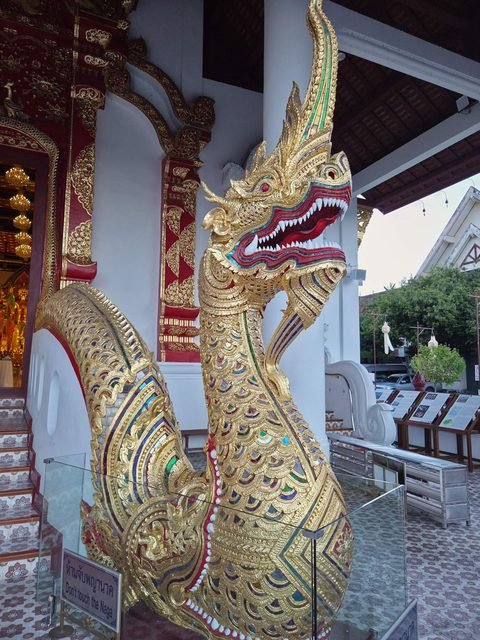 A gilded naga with coloured glass scales and an open red mouth at the stair of the Wihan Luang, behind a small 'Don't touch the Naga' sign