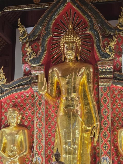 A tall gilded standing Buddha with the right hand raised, against a red patterned backdrop, a smaller gilded disciple figure on the left