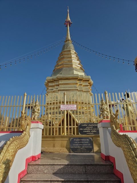 The gilded bell-shaped chedi behind a golden fence, at the top of a short stair with naga balustrades; plaques stand at the foot