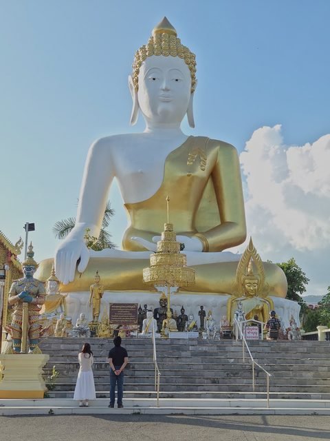 The big seated Buddha, white with a gilded robe, seen from below above a wide stair; two visitors stand at the foot of the steps, looking up