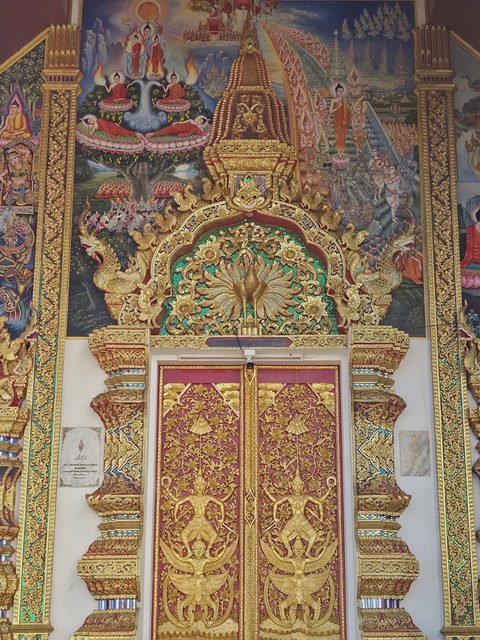 The doorway of the 1994 hall: carved and gilded doors under an arch of green mosaic with a golden peacock, between two painted scenes from the Buddha's life