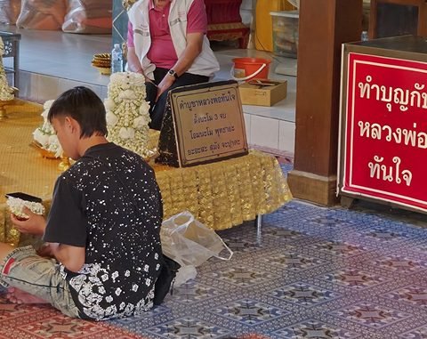 A worshipper sitting cross-legged on the tiled floor, seen from behind, holds a bowl of jasmine before a gilded platform; beside it, an engraved Thai plaque and a red donation box
