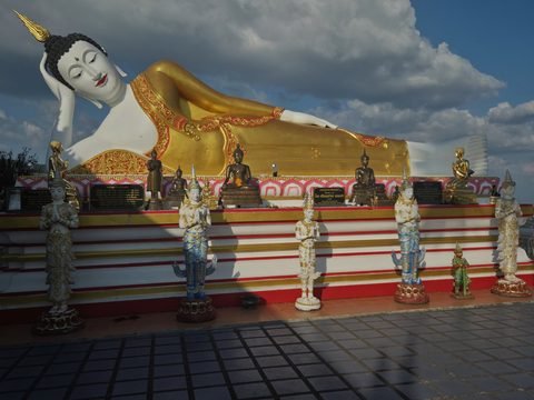 A reclining Buddha with a white face and a gilded robe on a terrace, small praying figures lined up in front of it