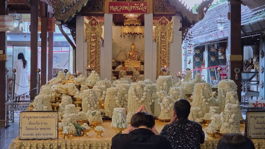 A small gilded Buddha in a shrine of red and gold pillars; in front, a platform covered with cones of white jasmine garlands; two men, seen from behind, pray with joined hands