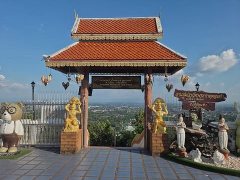 A gate with a tiled roof opening onto the plain, signed Doikham Panorama View Point, with two gilded figures carrying baskets and a large teddy bear in glasses