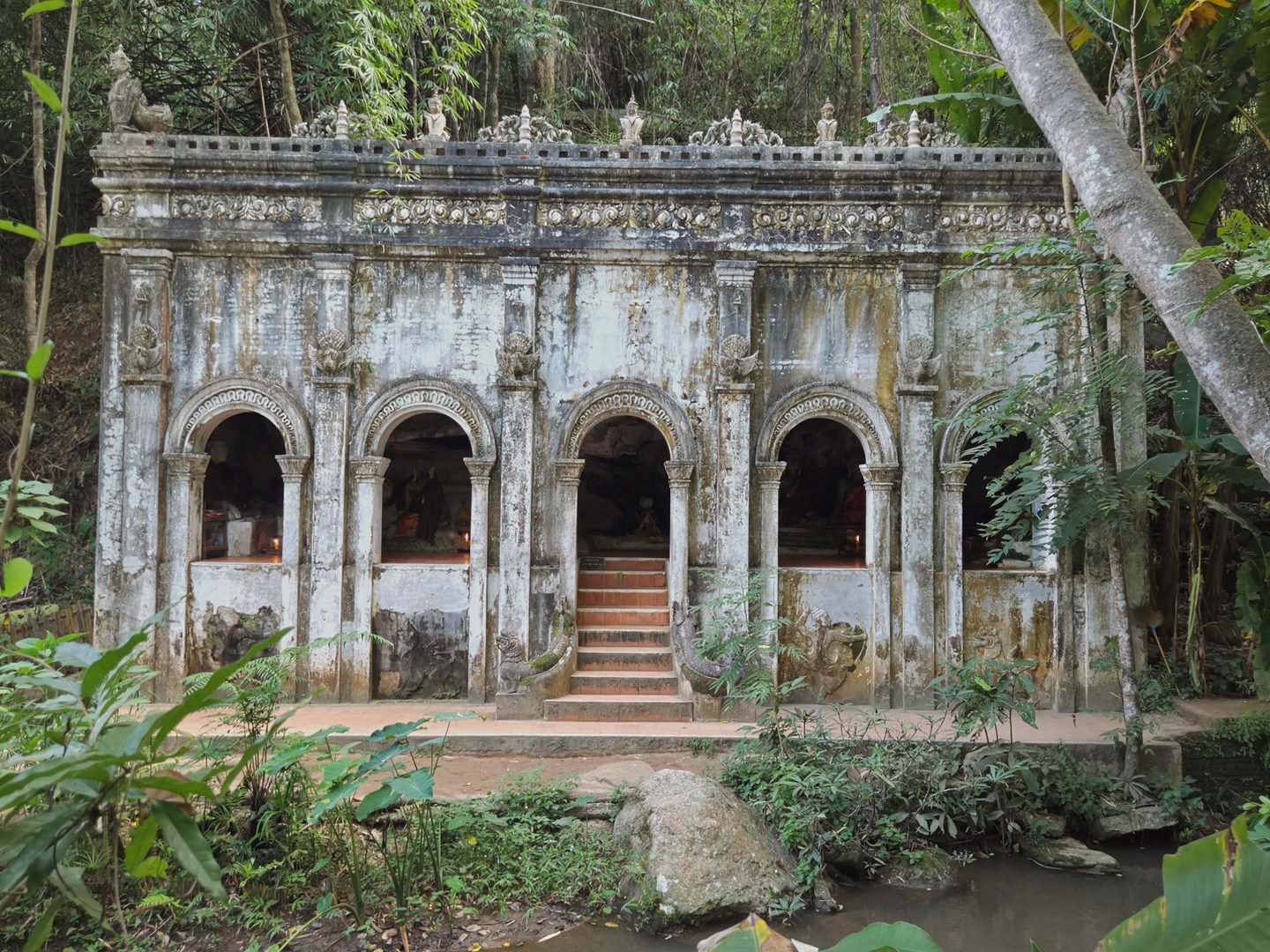 Un sanctuaire blanc à cinq arcades surmonté d'une balustrade de petites figures, ses murs striés d'humidité, en pleine forêt au-dessus d'un ruisseau rocheux
