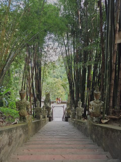 Un long escalier de pierre qui descend sous de hauts bambous, bordé de part et d'autre de petites figures de gardiens assis, vers une terrasse dallée
