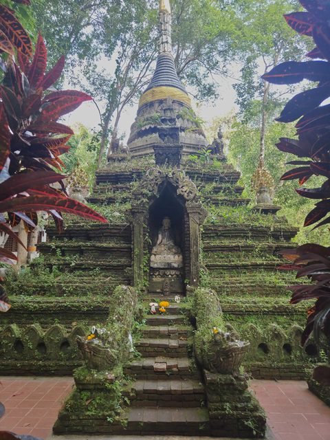 La face d'un chedi moussu : un escalier de pierre bordé de rampes à tête de serpent monte vers une niche abritant un Bouddha assis, sous une cloche sombre et une flèche dorée
