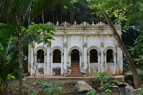 A weathered white temple hall with arched openings and red steps, surrounded by forest, with rocks and water in the foreground
