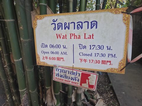 Un panneau « Wat Pha Lat » en thaï et en anglais, ouverture à 6 h, fermeture à 17 h 30, et un plus petit qui demande le silence, accrochés à des bambous