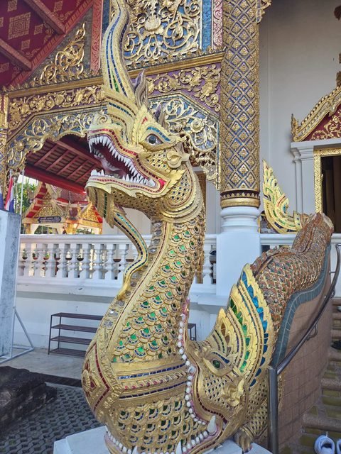 A gilded naga serpent with an open, toothed mouth rising from the jaws of a makara at the foot of the Wihan Luang stairs, its scales inlaid with coloured glass