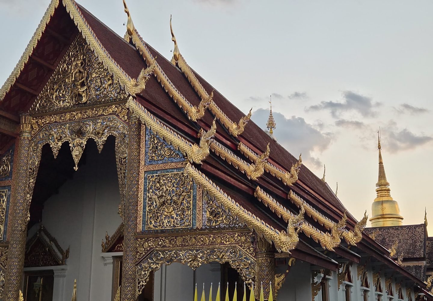 The gilded gables and stepped roofs of the Wihan Luang at dusk, with the gold spire of the chedi rising behind them on the right