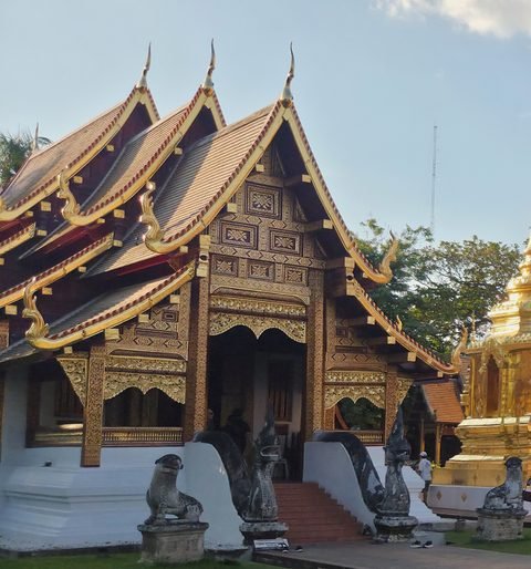 The front of the Wihan Lai Kham: stepped roofs edged in gold over a carved and gilded wooden gable, a white base, and naga stair rails flanked by stone guardian figures